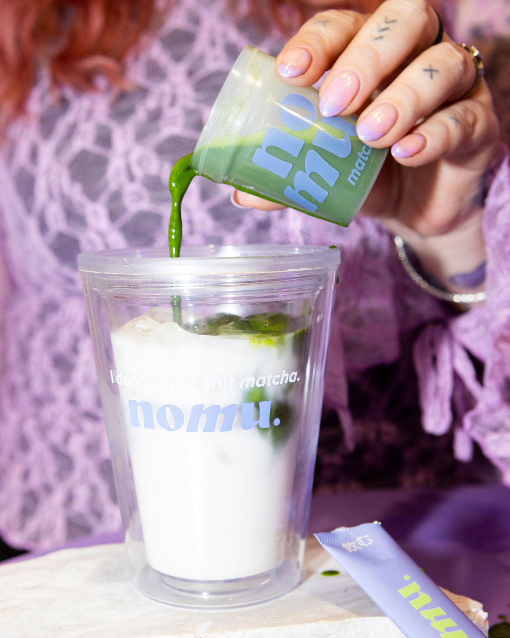 Person pouring matcha into a clear cup labeled 'nomu' with a green packet.