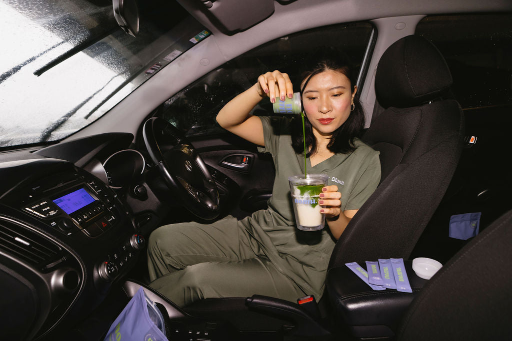 Woman sitting inside a car holding a drink and a straw, with various items on the car's interior.